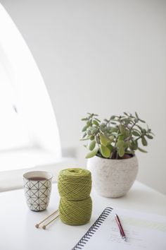 a white table topped with a plant and two cups filled with yarn next to a spiral notebook