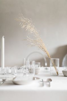 a white table topped with lots of glasses and plates next to a tall vase filled with dry grass