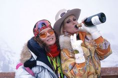 two women wearing winter clothes and hats, one holding a wine bottle while the other holds a camera