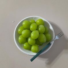 a white bowl filled with green grapes next to a blue fork on a beige surface