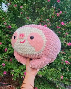 a hand holding up a pink and white crocheted stuffed animal in front of flowers