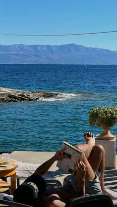 a man reading a book while sitting on a deck next to the ocean