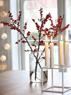 three candles are sitting on a table near a vase with red berries and branches in it