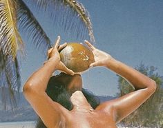 a naked woman holding a coconut in front of her face and palm tree on the beach