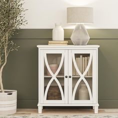 a white cabinet with glass doors next to a potted olive tree and a lamp