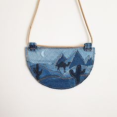 a denim purse hanging from a rope with cactus and mountains in the background on a white wall