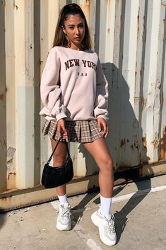 a young woman standing in front of a wall wearing a new york sweatshirt and plaid skirt