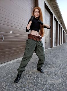 a woman with red hair and black gloves posing in front of a garage door wearing green cargo pants