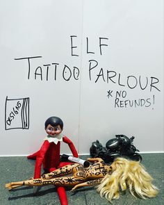 an elf is sitting next to a tattoo parlor sign and two dolls are lying on the ground