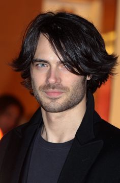 a man with long hair and blue eyes looks at the camera while wearing a black suit