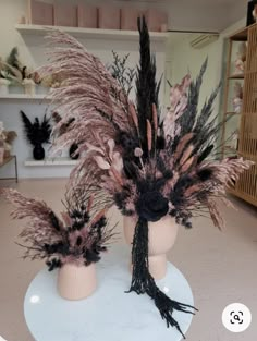 two vases with dried flowers and feathers on a table in a room filled with shelves