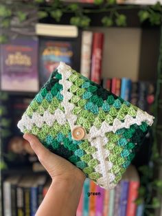 a hand holding up a green and white crocheted purse in front of a bookshelf