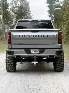 the back end of a silver chevrolet truck parked on a gravel road with trees in the background