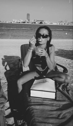 a woman sitting in a chair on the beach with a book and sunglasses around her neck