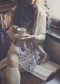 a woman sitting in a chair holding a cup and saucer while reading a book