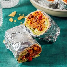 two burritos wrapped in aluminum foil on a blue tablecloth with other food items