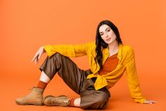 a woman is sitting on the floor wearing brown pants and a yellow shirt with an orange background