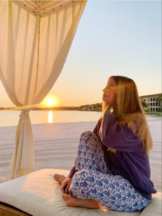 a woman sitting on top of a bed next to the ocean at sunset with her hair blowing in the wind