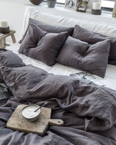 an unmade bed with grey linens in a white room next to a window