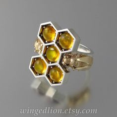 "HONEY BEES silver and 14k gold cocktail ring with Amber. The art of beekeeping goes back to ancient times. Over the centuries, bees and honey have been symbols of love, family, sweetness, and delight. HONEY BEES ring features a sterling silver honeycomb with cells filled with five natural 6mm amber cabochons. On the shank, flanking the honeycomb, there are two beautiful small honey bees made of 14k yellow gold. The shank is tapered from 6.8mm on top to 3.9mm on the bottom. On the inside of the Honeycomb Jewelry, Bee Ring, Gold Cocktail Ring, Gold Cocktail, Honey Bees, Themed Jewelry, 14k Gold Ring, Bee Keeping, Artistic Jewelry