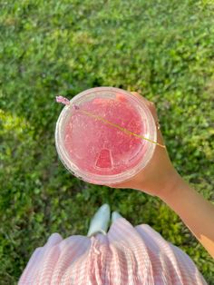 a person holding up a pink drink in their hand on the grass, looking down at it