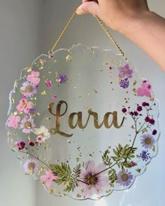 a hand holding a glass sign with flowers on it that says lara in gold lettering