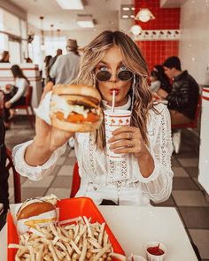 a woman sitting at a table with a sandwich and drink in her hand while holding a straw up to her mouth