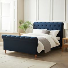 a bed with white sheets and blue headboard in a living room next to a window