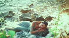 a woman in a blue dress is laying on the rocks by the water's edge