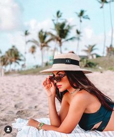 a woman laying on top of a beach wearing a hat and sunglasses with palm trees in the background