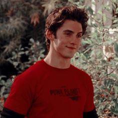 a young man in a red shirt standing next to some bushes and trees with his hands on his hips