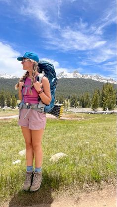 a woman standing in the grass with a backpack on her back and mountains in the background