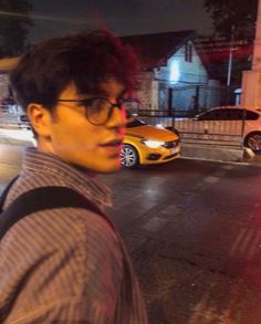 a man with glasses is standing on the street at night in front of some parked cars