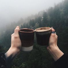 two people holding up coffee cups in the air with trees in the background on a foggy day