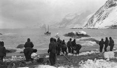 black and white photograph of people standing on the beach with boats in the water behind them