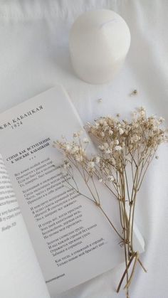 an open book next to some flowers on a white tablecloth with a vase and ball