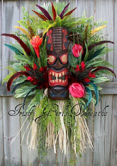 a tiki mask is adorned with tropical foliage and flowers in front of a wooden fence