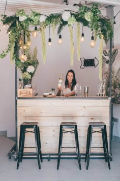 a woman sitting at a bar with three stools in front of it and greenery hanging from the ceiling