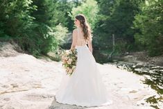 a woman in a wedding dress is standing on the rocks near water and holding a bouquet