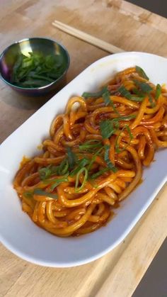 a white plate topped with noodles and sauce next to a bowl of greens on a wooden table