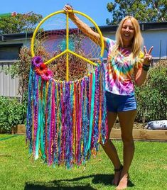 a woman standing in the grass holding up a rainbow colored dream catcher made out of yarn