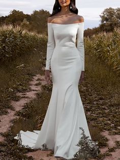 a woman in a white dress standing on a dirt path with grass and bushes behind her