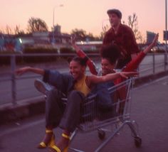 three people sitting in a shopping cart on the side of the road with their arms up