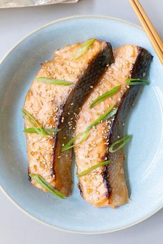 two pieces of fish on a blue plate with chopsticks next to it and another dish in the background