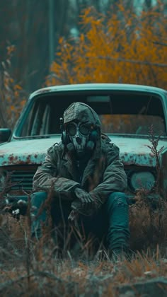 a man in gas mask sitting on the ground next to an old car with trees and bushes behind him