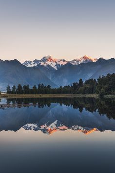 the mountains are reflected in the still water of this lake at sunset, with trees and bushes on either side