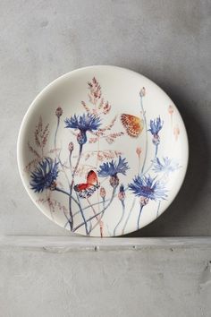 a white plate with blue flowers and birds painted on it sitting on a shelf next to a gray wall