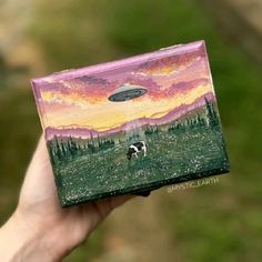 a hand holding up a painted box with a dog in the field and an alien hovering above it