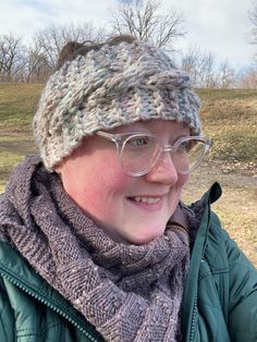 a woman wearing glasses and a knitted hat smiles at the camera with trees in the background