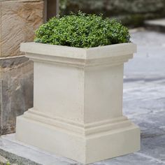 a white planter sitting on the sidewalk next to a stone wall with a green bush in it